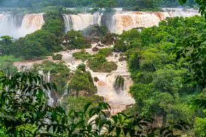 Imagen de una vista panorámica de las Cataratas del Iguazú en todo su esplendor, con el arco iris formado por la neblina de agua, la selva circundante y las pasarelas llenas de turistas en el primer plano.